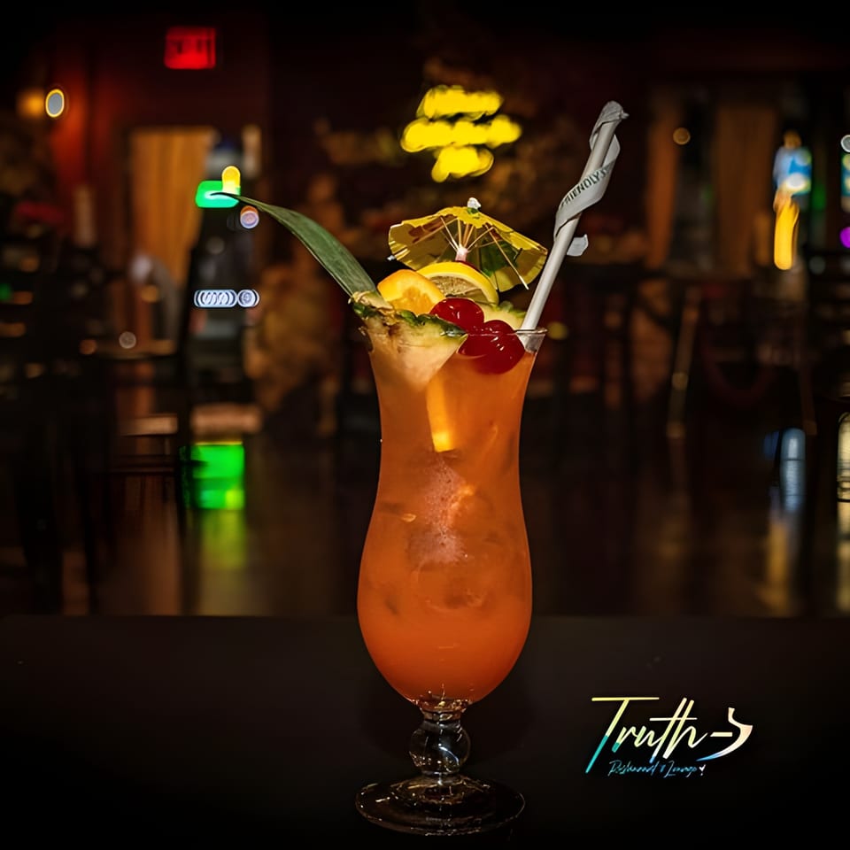 A vibrant Caribbean cocktail, garnished with fruit and a small umbrella, sits on a dark bar.