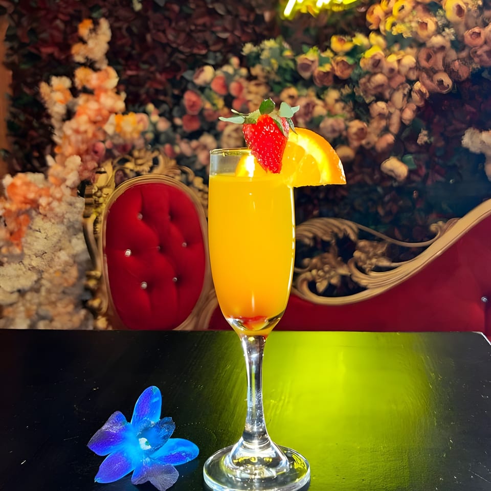 A mimosa with strawberry and orange garnish sits on a table with a blue orchid and red velvet chairs.